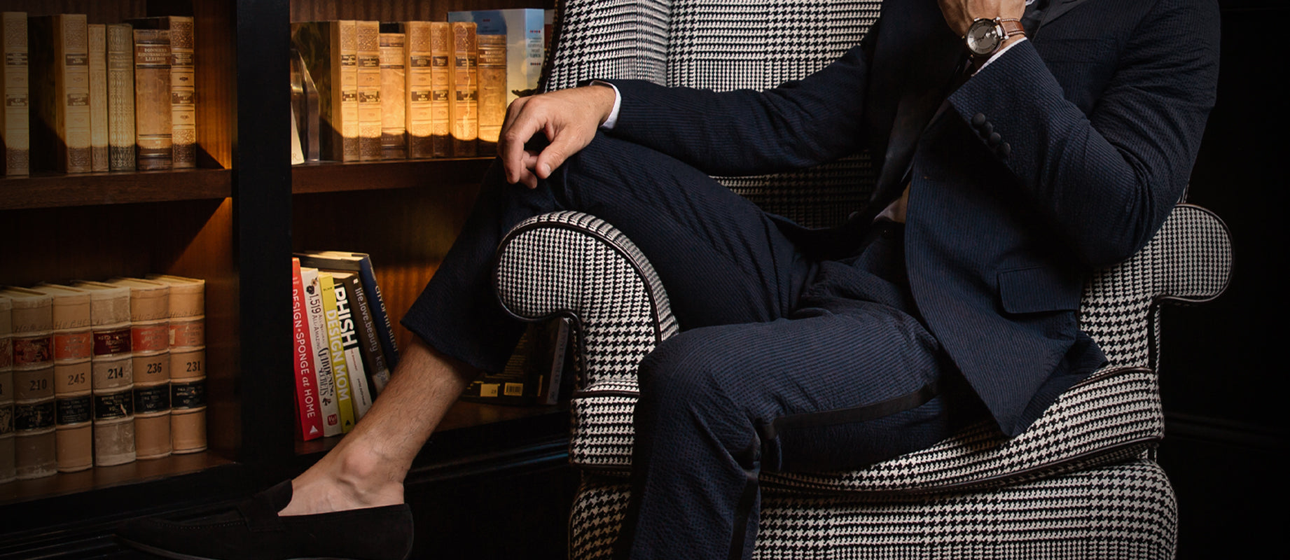 Man wearing a navy seersucker tuxedo and loafers sitting on a houndstooth arm chair in a sophisticated reading room.