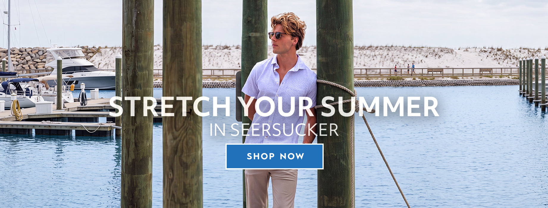 A stands on a dock by the water wearing a light blue button-up seersucker shirt and khaki pants, with large wooden posts framing the scene. White text across the center reads 'STRETCH YOUR SUMMER IN SEESUCKER' with a blue 'SHOP NOW' button below.