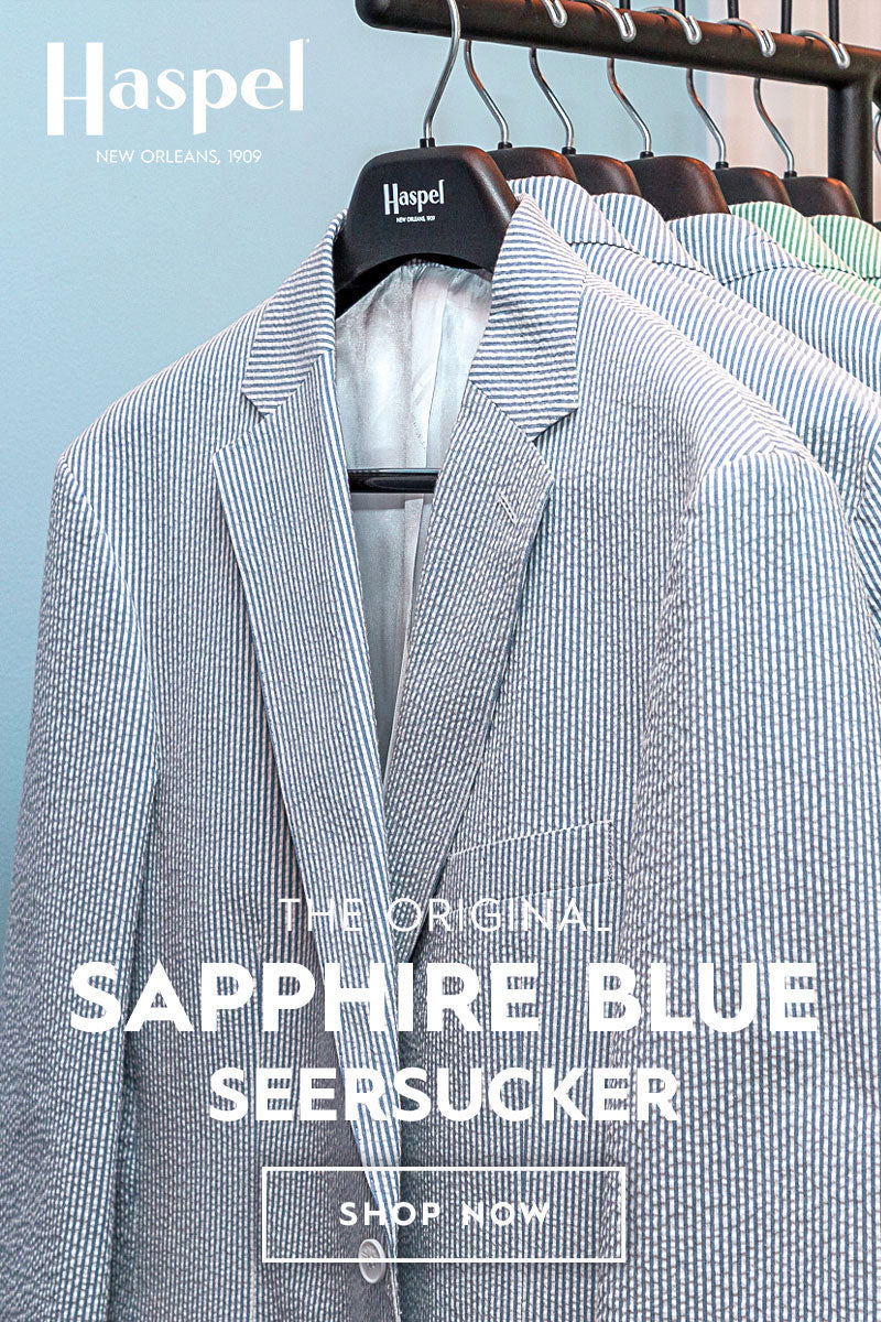 A close-up of a tailored blazer on a hanger with overlaid text reading 'The Original Sapphire Blue Seersucker' and a 'Shop Now' button. The blazer has a fine blue-and-white seersucker stripe pattern, and other hangers with similar garments are visible behind it against a pale blue background.